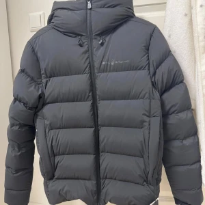 Svart dunjacka från Sail Racing - Snygg svart dunjacka från Sail Racing med huva och dragkedja. Perfekt för kyliga dagar med sin quiltade design som håller värmen. Jackan har en stilren look och är idealisk för både stad och natur. Tagsen kvar 