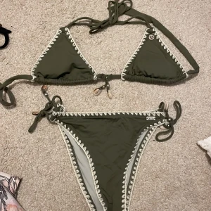 Olivgrön bikini med snäckdetaljer - Snygg olivgrön bikini från Banana moon med vita spetsdetaljer och snäckor vid knytningarna. Toppen har trekantsform och knyts i nacken och ryggen. Nypris 800 kr