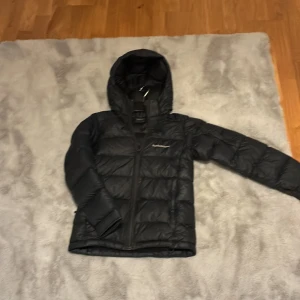 Svart dunjacka från Peak Performance - Säljer en svart dunjacka från Peak Performance. Jackan har en huva och dragkedja framtill. Perfekt för kyliga dagar med sin vadderade design.