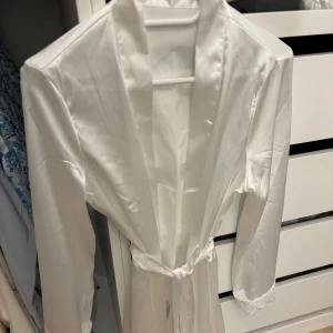 Vit sidenmorgonrock med spetsdetaljer - Elegant vit morgonrock i siden med långa ärmar och spetsdetaljer vid ärmslut. Den har ett knytband i midjan för en justerbar passform. Perfekt för en lyxig känsla hemma. Aldrig använd. 