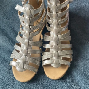 Silvriga sandaler med flätad detalj - Snygga silvriga sandaler med flera remmar och en flätad detalj. De har en öppen tå och justerbar rem runt ankeln. Perfekta för sommarens alla tillfällen.