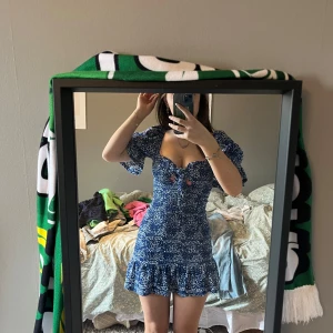 Blå blommig klänning - Charmig blå klänning med blommigt mönster och volangdetaljer. Klänningen har korta ärmar och en v-ringad design med knappar framtill. Perfekt för sommarens alla tillfällen.