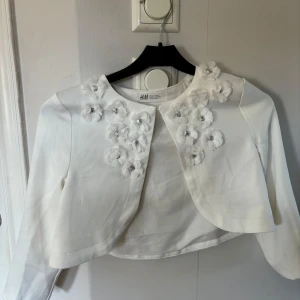Vit bolerotopp från H&M - Säljer en söt vit bolerotopp från H&M med blommiga detaljer och pärlor framtill. Toppen har långa ärmar och en rund halsringning. Perfekt för speciella tillfällen! 🌸Skriv om ni undrar något!💗
