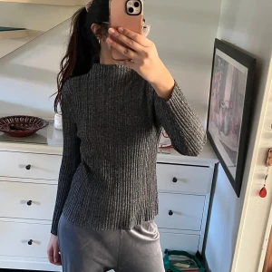 Grå ribbad tröja - Säljer en stilren grå ribbad tröja med långa ärmar och hög krage. Perfekt för kyligare dagar och lätt att matcha med olika outfits. Passar både till vardags och mer uppklädda tillfällen.
