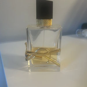 Parfym YSL - Elegant parfymflaska med en fyrkantig design och gulddetaljer. Flaskan har en svart kork med guldfärgad textur vid basen. En gyllene logotyp pryder framsidan, vilket ger en lyxig känsla. Nypris är 1400, säljer för 500