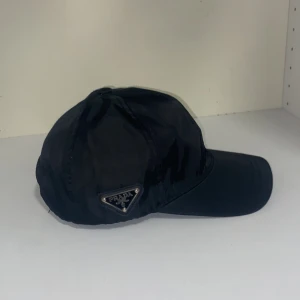 Svart keps från Prada - Snygg svart keps från Prada i bomull med justerbart spänne baktill. Klassisk design med Pradas logotyp på sidan och broderad text på baksidan. Perfekt för en stilren look. Priset kan diskuteras. Tar emot byten också bara att komma med förslag!!