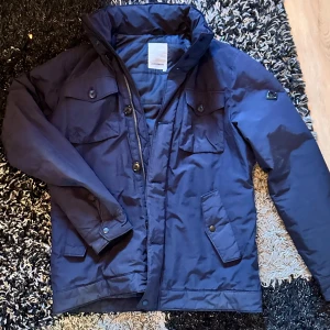 Blå jacka från J.Lindeberg - Snygg blå jacka från J.Lindeberg med flera fickor och knappar. Jackan har en dragkedja och är perfekt för kyligare dagar. Den har en stilren design med långa ärmar och en hög krage.