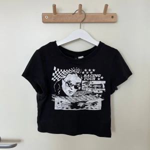 Svart croppad t-shirt - Säljer en svart croppad t-shirt från H&M Divided med ett coolt tryck av en person och texten 'Racing Tour'! Använd endast en gång ❤️