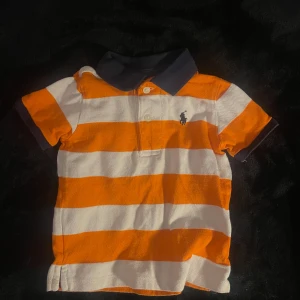 Orange och vit randig pikétröja från Ralph Lauren - Säljer en snygg orange och vit randig pikétröja från Ralph Lauren. Tröjan har en klassisk krage i mörkblått och korta ärmar. Perfekt för en stilren look! 🧡