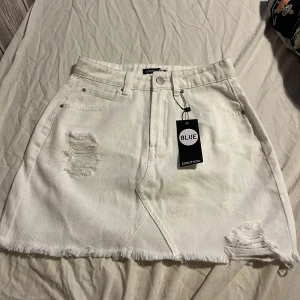 Jeanskjol - Snygg vit jeanskjol från Boohoo med slitna detaljer. Kjolen har en klassisk ficksdesign och stängs med knapp och dragkedja. Perfekt för en snygg look!