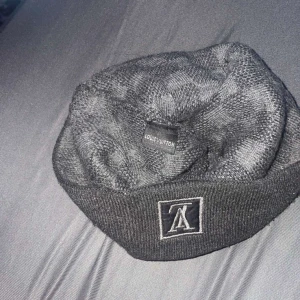 Grå rutig mössa från Louis Vuitton - Snygg grå mössa från Louis Vuitton med ett klassiskt rutigt mönster. Mössan har en uppvikt kant och en diskret logotyp framtill. Perfekt för att hålla stilen i kallare väder.