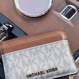 Plånbok från Michael Kors - Snygg och stilren plånbok från Michael Kors i beige och brun färg. Den har flera kortfack och är perfekt för att hålla ordning på dina kort. Plånboken är kompakt och lätt att bära med sig.