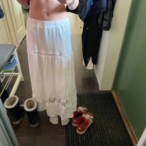Vit långkjol med spetsdetaljer - Säljer en vacker vit långkjol med spetsdetaljer. Kjolen har en elastisk midja och fina spetsinslag som ger en romantisk känsla. Perfekt för varma dagar eller sommarens fester. Dock en liten fläck som inte syns på bilderna men be om bild på den så skickar jag🩷