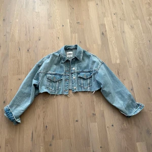 Blå croppad jeansjacka - Säljer en snygg blå croppad jeansjacka från Zara. Jackan har en klassisk krage, knappar framtill och två bröstfickor med lock. Den har en rå kant nedtill för en trendig look. Perfekt för att lägga till en cool touch till din outfit.