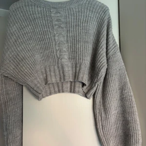 Grå stickad tröja från NA-KD - Säljer en snygg grå stickad tröja från NA-KD. Tröjan har ett flätat mönster framtill och är croppad med långa ärmar. Perfekt för kyligare dagar!