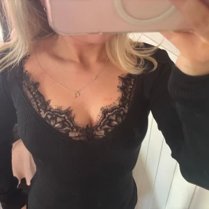 Svart ribbad topp med spetsdetaljer - Snygg svart ribbad topp med v-ringning och spetsdetaljer vid halsen. Perfekt för en stilren look. Långärmad och figurnära passform.