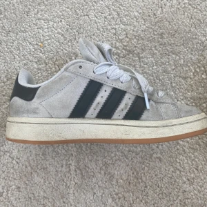 Grå Beige Adidas Campus  - Säljer ett par grå Adidas Campus sneakers med svarta ränder. De är ganska smutsiga därav priset men annars inga skador 
