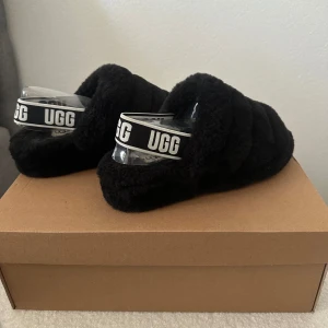 Svarta fluffiga tofflor från UGG - Säljer ett par svarta fluffiga tofflor från UGG med öppen tå och elastiskt band med logga. Perfekta för att hålla fötterna varma och bekväma utomhus.