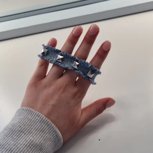 Jeansarmband med nitar - Snyggt armband i jeansmaterial med silverfärgade nitar. Armbandet har en rå kant och stängs med en tryckknapp. Perfekt för att ge en edgy touch till din outfit.🩷 armbandet är 18,5 cm