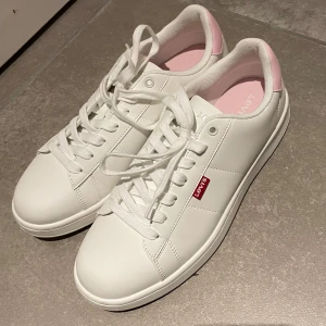 Vita sneakers från Levi's - Snygga vita sneakers från Levi's med rosa detaljer på hälen. Skorna har klassisk snörning och en rund tå. Perfekta för en stilren look med en touch av färg. Använda en gång.