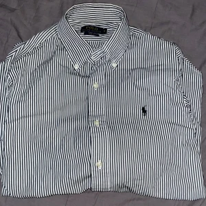 Randig skjorta från Ralph Lauren - Snygg svartvit randig skjorta från Ralph Lauren i slim fit. Skjortan har långa ärmar och knappar framtill. Perfekt för en stilren look med klassisk design och ikonisk logga på bröstet. I nyskick utan några fläckar. Pris kan fiskuteras