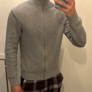 Grå stickad kofta med dragkedja - Säljer en stilren grå stickad kofta med dragkedja. Perfekt för kyliga dagar med sin höga krage och långa ärmar. Enkel att matcha med olika outfits.