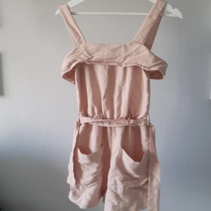 Ljusrosa jumpsuit - Ljusrosa byxdress med fickor och breda axelband. Jumpsuiten har en dragkedja bak och elastisk midja för en bekväm passform med knytbälte. Axelbanden kan justeras i längd i 3 lägen. Små skavanker som en lös tråd och några små fläckar (som säkert går bort med lite fläckborttagning), därav priset 💗