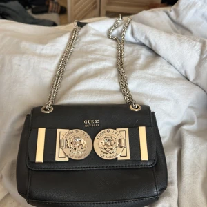 Svart axelväska från Guess - Snygg svart axelväska från Guess med kedjeaxelrem och dekorativa gulddetaljer. Väskan har en elegant design med Guess-logga och ett praktiskt innerfack med dragkedja. Perfekt för att bära dina nödvändigheter med stil.