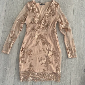 Champagnefärgad paljettklänning från Boohoo - Snygg champagnefärgad klänning från Boohoo med glittrande paljetter över hela plagget. Klänningen har en v-ringad design och långa ärmar i ett transparent material. Perfekt för en festlig kväll!