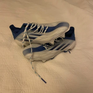 Vita och blå fotbollsskor från Adidas - Helt oanvända men en liten fläck (går nog att ta bort)!Säljer ett par vita och blå fotbollsskor från Adidas med snörning och dobbar för optimalt grepp på planen. Perfekta för fotbollsträning och matcher. Skorna har en stilren design med klassiska Adidas-ränder.