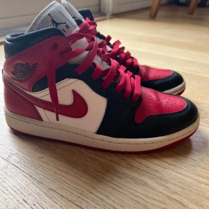 Nike Air Jordans 1 mids i rött, svart och vitt - Snygga Nike Air Jordans i en klassisk färgkombination av rött, svart och vitt. Skorna har en ikonisk design med snörning och en robust röd sula. Använda men i mycket bra skick. 