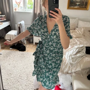 Grön blommig omlottklänning - Säljer en söt grön omlottklänning från Aachoe med vita blommor. Klänningen har korta ärmar och en knytning i midjan som ger en fin passform. Perfekt för sommarens alla tillfällen! 🌸Storleken är S men eftersom den har knytningen kan man justera den så skulle säga att den passar XS-M🥰