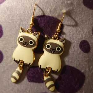 Söta kattörhängen i guld och vit - Charmiga örhängen med kattmotiv i guld och vit. De har en lekfull design med stora ögon och randig svans, perfekt för kattälskare. Hängena är fästa på krokar för enkel användning.