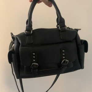 Svart axelväska med nitar - Snygg svart axelväska i syntetmaterial med coola nitar och dragkedja. Väskan har både handtag och en justerbar axelrem för flexibilitet. Perfekt för vår och sommar, säljs pga användning helt nytt skick, pris diskuter bart