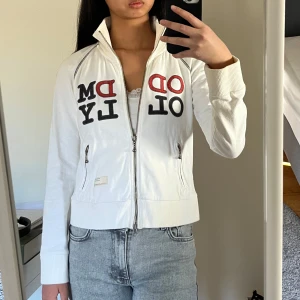 Vit Odd Molly kofta med text - Säljer en vit kofta med dragkedja och Odd Molly tryck i rött och svart på framsidan. Jackan har långa ärmar och ribbade muddar vid ärmslut och nederkant. 