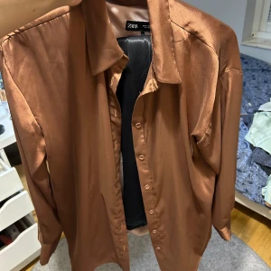 Brun satinliknande skjorta från LCW Vision - Säljer en brun skjorta i glansigt satinliknande material från LCW Vision. Skjortan har klassisk krage, knappar framtill och långa ärmar. Perfekt för dig som vill ha en stilren och trendig look.