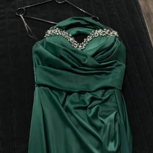 Grön långklänning med pärldetaljer - Säljer en elegant grön långklänning med vacker drapering och pärldetaljer längs halsringningen. Klänningen har en omlottliknande överdel och är perfekt för speciella tillfällen.