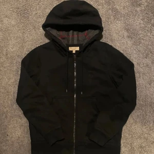Burberry Hoodie - Säljer en svart hoodiejacka från Burberry med dragkedja och huva. Insidan har ett klassiskt grått och rött rutigt foder. Jackan har långa ärmar och två fickor framtill. Perfekt för en stilren och avslappnad look