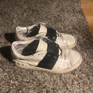 Valentino Open sneakers med svart band - Säljer ett par vita Valentino Opens. Perfekta för dig som gillar stilrena och exklusiva sneakers inför sommaren! Skorna är i okej skick samt har två hål under båda skor men gör inget under sommaren. Kontakta gärna om ni har frågor samt funderingar. Priset går att diskuteras vid snabbaffär!