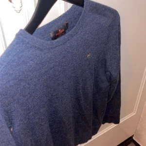 Blå stickad tröja från Morris - Säljer en klassisk blå stickad tröja från Morris med rund halsringning och diskret broderad logga på bröstet. Tröjan har långa ärmar och ribbade muddar vid ärmslut och nederkant. Perfekt för lager-på-lager-stil.