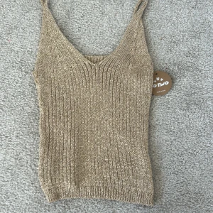 Beige ribbad linnetopp från Motivo - Säljer en beige ribbad linnetopp från Motivo med smala axelband och v-ringning. Toppen har en glittrig finish och är perfekt för sommaren. Snygg att styla till både jeans och kjol!