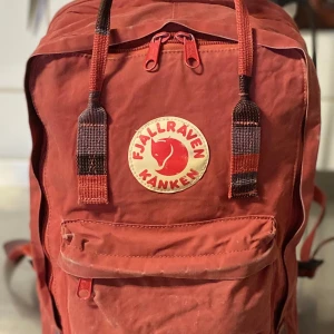 Röd ryggsäck från Fjällräven Kånken - Säljer en klassisk röd Fjällräven Kånken ryggsäck med det ikoniska runda märket framtill. Väskan har justerbara axelremmar, dragkedja upptill och en stor ytterficka med dragkedja. Perfekt för skola eller utflykter.Titta mer på bilden.