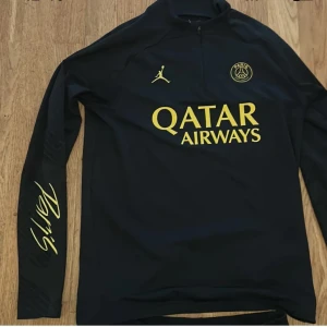 Svart långärmad PSG fotbollströja med gul text - Säljer en svart långärmad PSG träningströja med gul text och logga. Tröjan har halv dragkedja, Jordan-logga och Paris Saint-Germain-märke på bröstet samt Qatar Airways som sponsor. Snygg detalj med 'Paris' på ärmen.