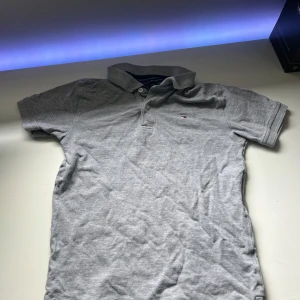 Grå pikétröja från Tommy Hilfiger - Klassisk grå pikétröja från Tommy Hilfiger med liten broderad logga på bröstet. Tröjan har korta ärmar, krage och knappar framtill. Perfekt för en clean och stilren look. Storlek 152-158