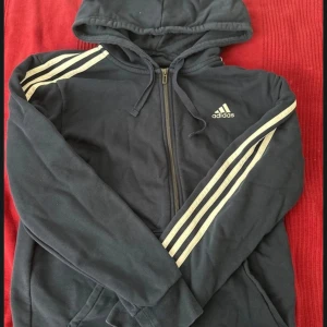 Mörkblå hoodie med dragkedja från Adidas - Mörkblå hoodie från Adidas med vit logga på bröstet och klassiska tre vita ränder längs axlarna och ärmarna. Jackan har huva med dragsko, dragkedja framtill och två fickor.