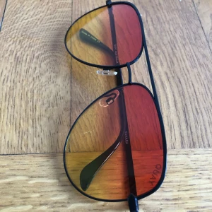 Quay solglasögon med gula och orange glas - Snygga pilotbågar från Quay med svarta skalmar och färgade glas i gult och orange. Metallbågarna ger en klassisk look och glasen har en cool gradient-effekt. Perfekta för att sticka ut i sommar.