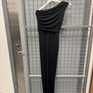 Svart one shoulder långklänning köpt på bubbleroom - Säljer en elegant svart långklänning från Zack London med one shoulder-design och draperad överdel. Klänningen har en hög slits framtill och smickrande passform, perfekt för speciella tillfällen. Storlek 8=xs