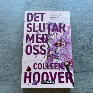 Det slutar med oss - Säljer boken 'Det slutar med oss' av Colleen Hoover. Omslaget är ljust rosa med lila text och blommotiv. Romanen är en gripande kärlekshistoria som utspelar sig i Boston och är skriven av en bästsäljande författare.