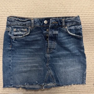 Jeanskjol från Gina Tricot  - Säljer en klassisk blå jeanskjol från Perfect Jeans med rå kant nertill och femficksmodell. Kjolen har knappgylf och tydliga sömmar i kontrastfärg. Perfekt för dig som gillar en avslappnad och tidlös stil.