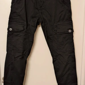 Svarta cargopants från Alpha Industries - Säljer ett par svarta cargopants från Alpha Industries med flera fickor och snygga detaljer. Byxorna har muddar vid bensluten och en röd etikett med logga. Perfekta för en streetwear-look.